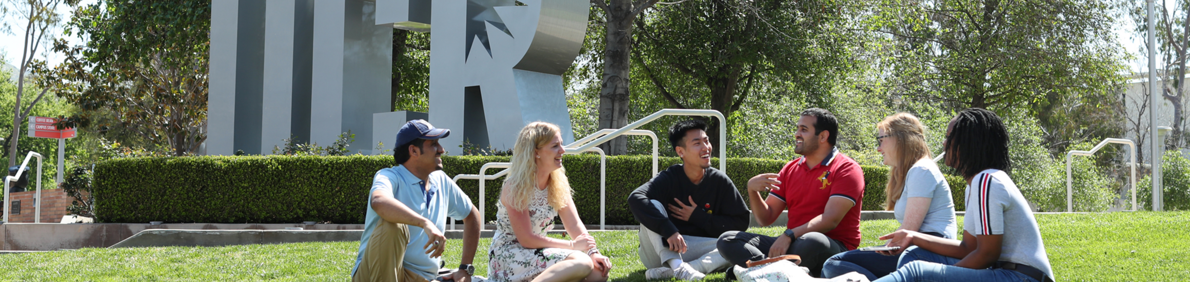 A group of students sit in a semi-circle and chat on the lawn in front of the UCR letter sculpture on the UC Riverside campus.